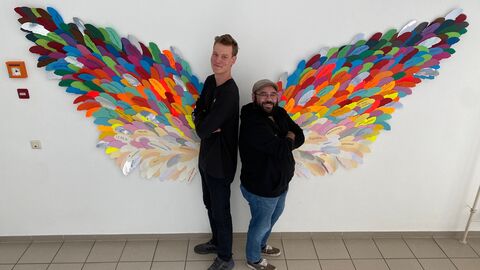 Ein großer und ein kleiner Mann stehen vor bunten Flügeln. Die bunten Flügel sind aus kleinen Zetteln zusammengefügt und an die Wand gebracht. 