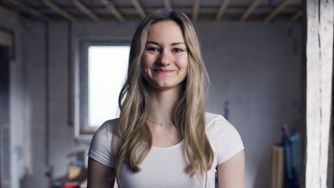 Lilly ist eine junge Frau mit langen blonden Haaren und steht in ihrem Eigenheim, das sie renoviert.